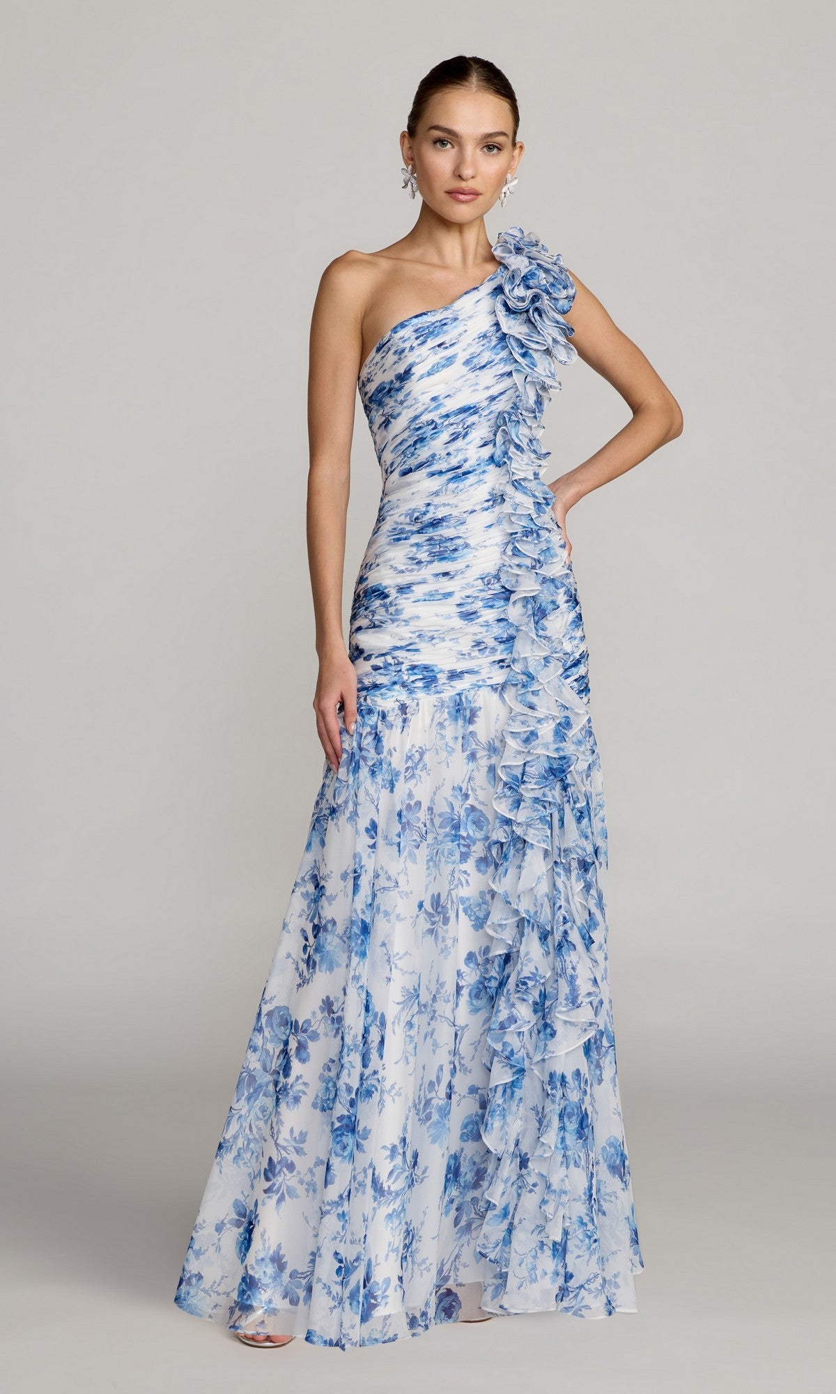 A woman wearing the Mac Duggal 68992 long formal dress in Blue Multi, featuring elegant one-shoulder styling with blue floral patterns and ruffled detailing, stands against a plain gray background.